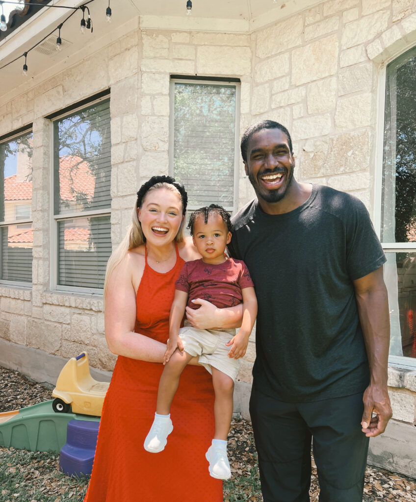 Iskra is holding her baby standing next to Philip, smiling in a casual family photo outside their home.