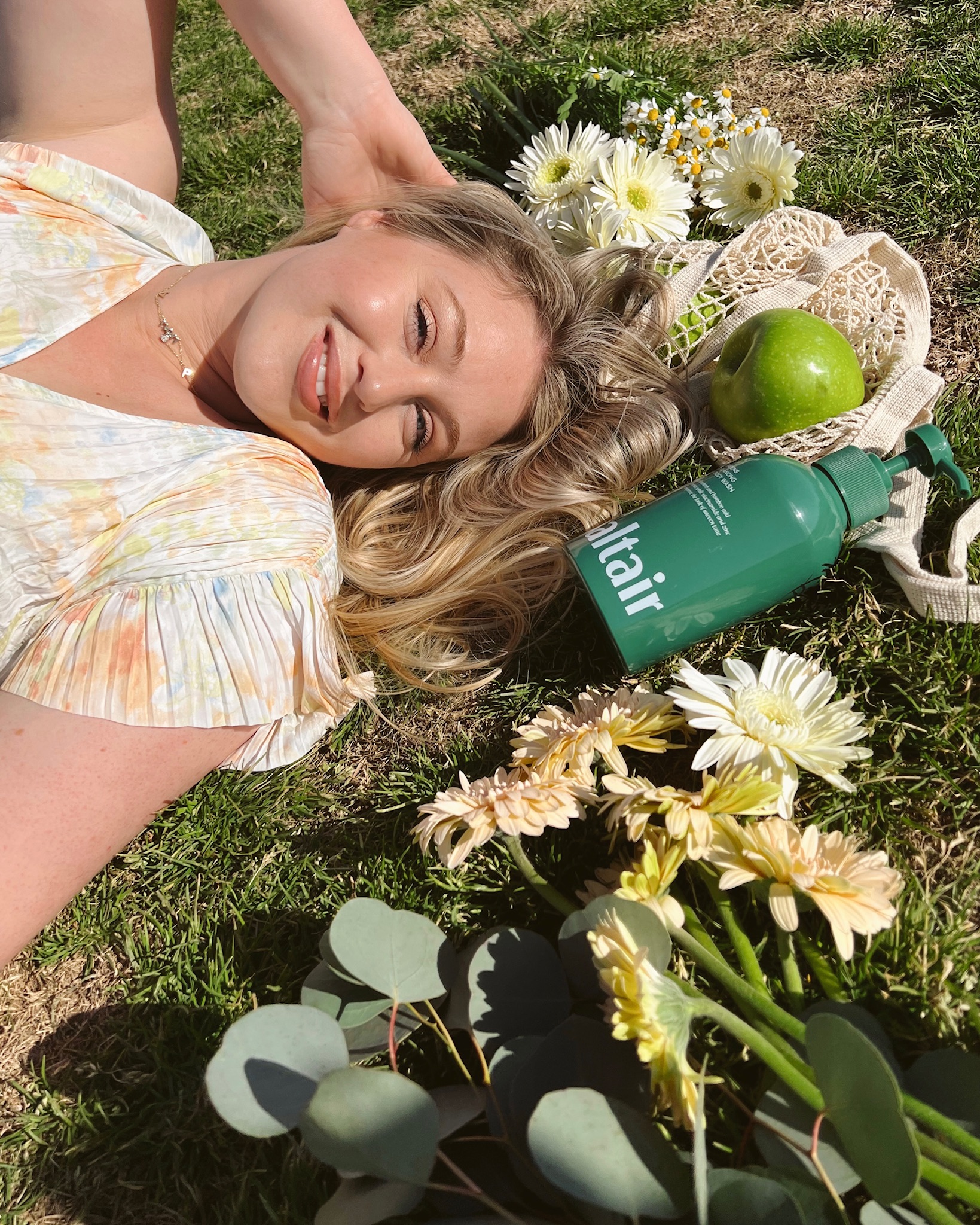 Iskra is laying down on the grass in a floral top with a Lush Greens Saltair body wash and some flowers surrounding her hair.