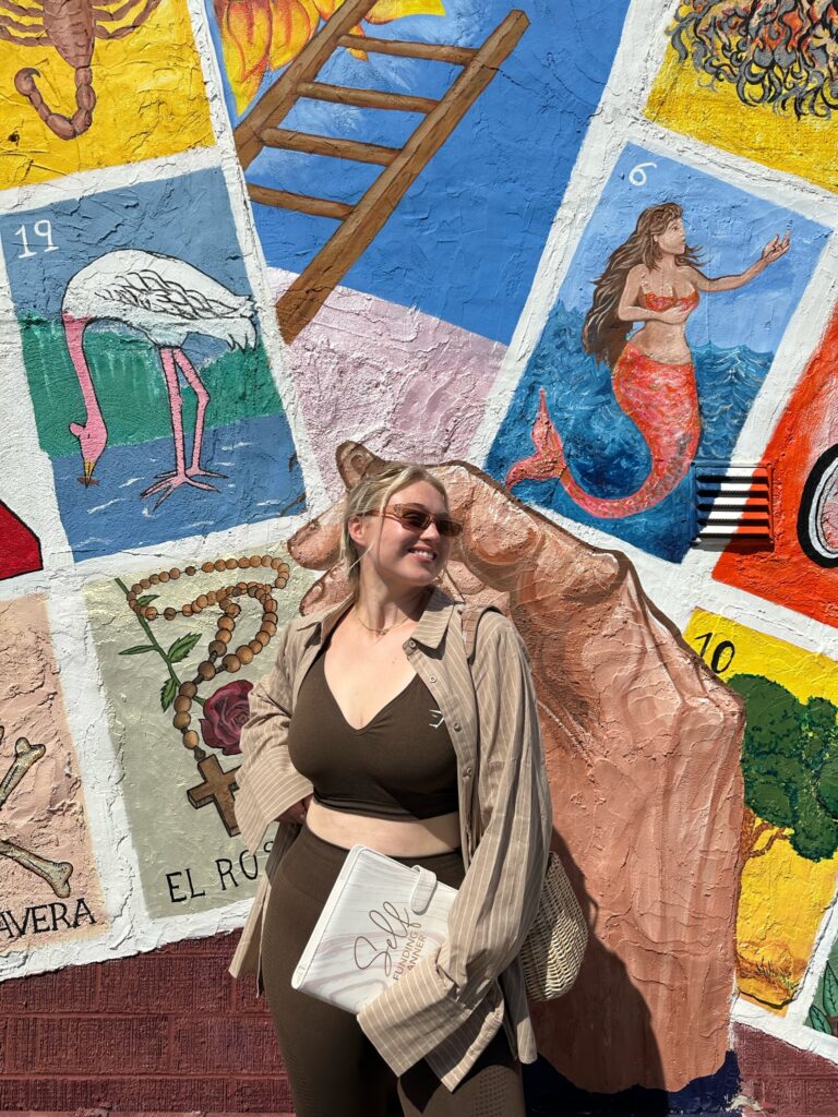 Iskra is holding her Self Funding Planner down in front of her, wearing a brown athleisure outfit and standing in front of a colorful mural with a Loteria card game theme.